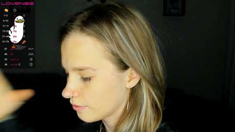 Media: Video of a young woman with light skin and straight blonde hair, wearing a black top, looking contemplative. Background shows a dimly lit room with a cartoon duck on a digital screen.