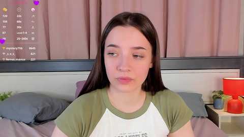 Media: Video of a young woman with straight black hair, wearing a green and white T-shirt, sitting on a bed with gray sheets and pillows. The background features a window with pink curtains and a lamp on a bedside table.