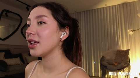 Media: Video of an Asian woman with straight, dark hair in a ponytail, wearing a white spaghetti-strap top and white earbuds, standing indoors. Background features a heart-shaped mirror, beige curtains, and a beige couch with a plush cat toy.