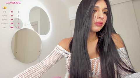 Media: Video of a transgender woman with long, straight black hair, wearing a white, off-the-shoulder fishnet top and silver elbow-length gloves, standing in a brightly lit room with a round mirror and calendar.