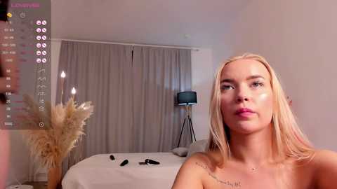 Media: Video of a topless, light-skinned, blonde woman with a tattoo on her shoulder, standing in a minimalist bedroom with beige curtains, a lamp, and a bed.