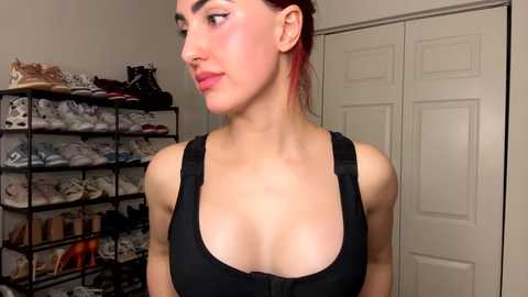 Media: Video of a young woman with fair skin, brown hair, wearing a black sports bra, standing in a cluttered room with shoe racks and closed doors.