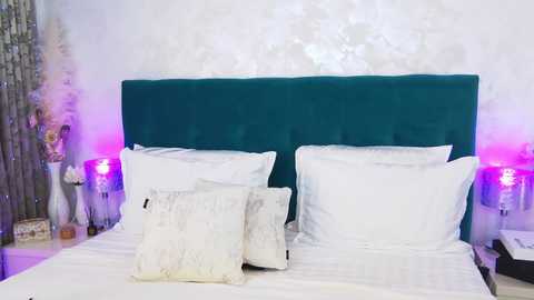 Media: Video of a modern bedroom with a teal tufted headboard, white bedding, and decorative pillows. Purple and white accent lights, and a vase with tall white flowers on the left.