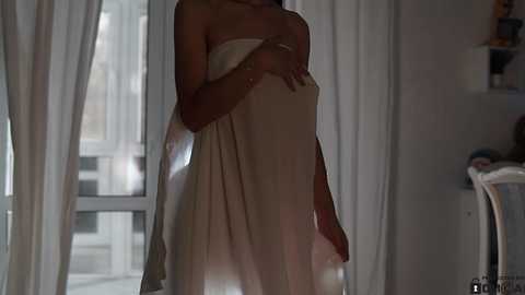 Media: Video of a pregnant woman in a white strapless dress, standing in a dimly lit room with white curtains and a window. She covers her belly with her hands.