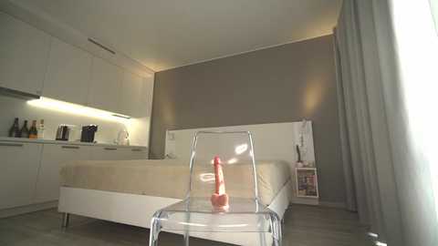 Media: Video of a minimalist bedroom with white furniture, beige bedspread, glass chair, and a red dildo on a glass table; soft natural light.