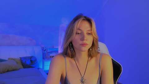 Media: Video of a young Caucasian woman with shoulder-length blonde hair, wearing a gray spaghetti-strap top, sitting on a black gaming chair under blue LED lights in a bedroom.