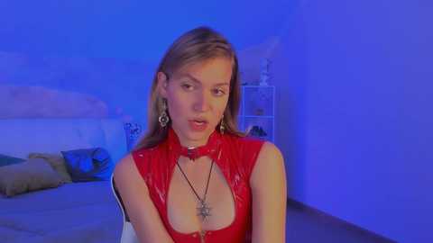 Media: Video of a Caucasian woman with light skin, blonde hair, wearing a red latex dress with a plunging neckline, standing in a dimly lit room with blue lighting, looking seductively.