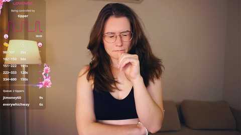 Media: A video of a fair-skinned woman with long brown hair, wearing glasses and a black tank top, contemplating with her finger to her lips, in a softly lit living room.