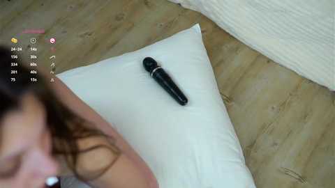 Media: Video of a woman lying on a white pillow, holding a black vibrator. She has long, wavy brown hair and is partially visible on the left side. The background shows a beige wooden floor.