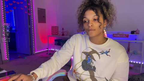 Media: Video of a young Black woman with curly hair, wearing a graphic sweatshirt, playing a keyboard in a dimly lit room with purple and blue LED lights.