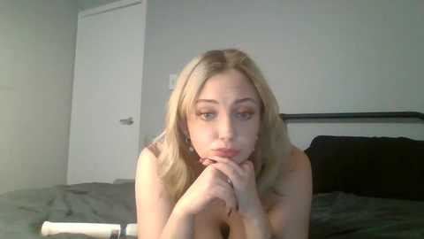 Media: Video of a young blonde woman with fair skin, resting on a bed with gray sheets. She's topless, gazing pensively with her chin on her hands. Background includes a white door and gray wall.