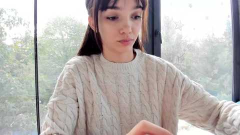 Media: Video of a young woman with straight brown hair, wearing a beige knitted sweater, standing by a large window with a blurred outdoor greenery view. She has a neutral expression.