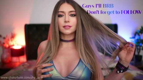 Media: Video of a young, light-skinned woman with long, straight blonde hair, wearing a low-cut, tie-dye top, holding her hair. She has a choker and a black wristband. Text overlays say \"Cums Fill BBB Don't for get to FOL!\