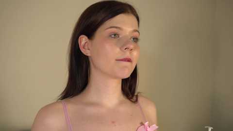 Media: Video of a young Caucasian woman with fair skin, straight brown hair, and a slender build, wearing a light pink spaghetti-strap top with a pink floral accessory. She stands against a plain beige wall, looking thoughtful.