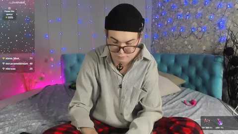 Media: Video of a young man with glasses and a black beanie, wearing a beige shirt, lying on a bed with a teal headboard, surrounded by fairy lights and pink and blue ambient lighting.