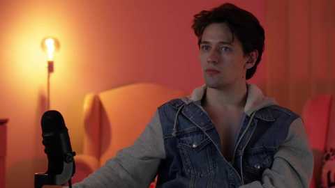 Media: Video of a young man with short brown hair, wearing a denim jacket, sitting in a dimly lit room with a warm orange glow and a lamp.