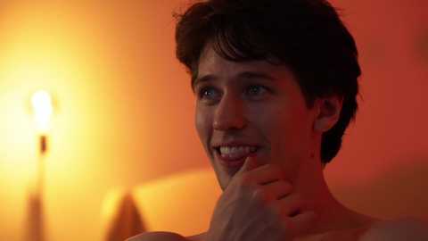 Media: Video of a smiling, shirtless young man with short dark hair, illuminated by a warm, orange-tinted lamp in a softly blurred background.