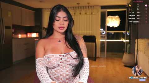 Media: Video of a Latina woman with long black hair, wearing a white, off-shoulder lace top, in a modern, dimly-lit living room with wooden floors.