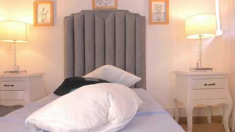 Media: Video of a neatly made bed with white pillows, gray tufted headboard, two white nightstands with lamps, framed art on beige walls, soft lighting.