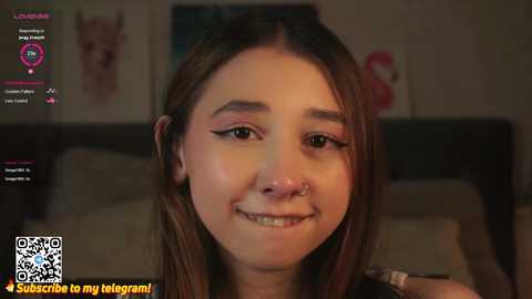 Media: A video of a young woman with fair skin, brown hair, and a nose ring, smiling in a dimly-lit bedroom with blurred background.