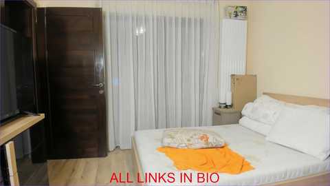 Media: Video of a minimalist bedroom with a single bed, white sheets, and orange blanket; a wooden door and white curtains; a flat-screen TV on the left. Text reads \"ALL LINKS IN BIO\".