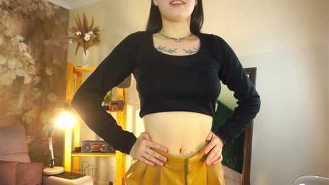 Media: Video of a young woman with light skin and black hair, wearing a black long-sleeve crop top, yellow high-waisted skirt, and floral tattoo on her chest. She stands confidently in a cozy living room with warm lighting.