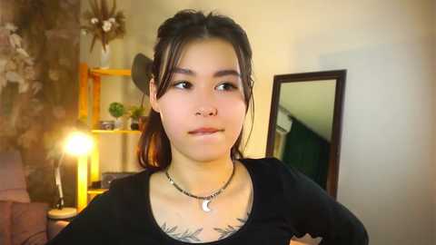 Media: Video of a young woman with medium skin tone, long dark hair, and a nose piercing, wearing a black top, standing indoors. She has tattoos on her chest and is smiling slightly.