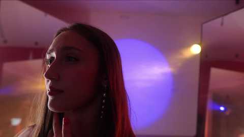 Media: Video of a woman with long brown hair, wearing a red dress, gazing introspectively in a dimly lit room with purple and yellow lights.