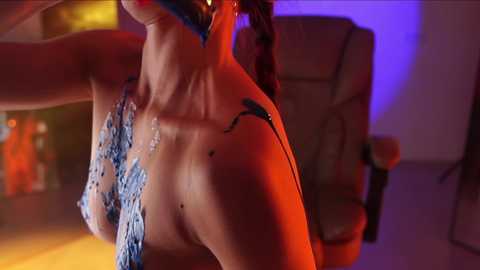 Media: Video of a slender, topless woman with light skin, wearing a blue sequined bra, standing in a dimly lit room with a plush chair and purple and red lighting.