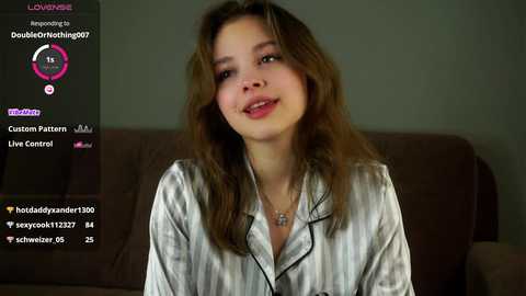 Media: Video of a young, light-skinned woman with wavy brown hair, wearing a silver satin robe, smiling against a brown couch. Digital interface with live stream and chat features in the background.