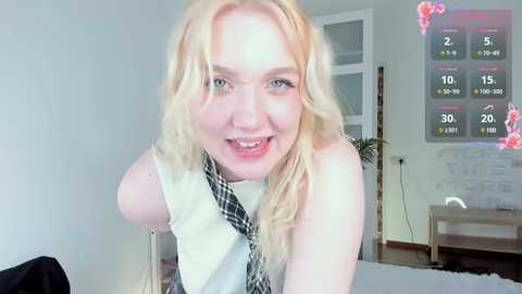 Media: Video of a smiling, light-skinned, blonde woman with wavy hair wearing a white sleeveless top and black-and-white patterned tie, in a bright, modern living room with a colorful digital clock and abstract wall art.