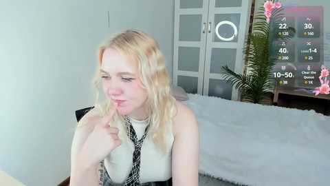 Media: A video of a young, fair-skinned blonde woman with wavy hair, wearing a white sleeveless dress and black tie, biting her finger, indoors, with a white bed, green plant, and digital clock in the background.