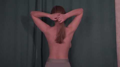 Media: A video of a topless woman with light skin and straight brown hair, facing away from the camera, arms raised, hands clasped behind her head, against a green curtain background.