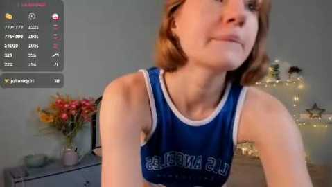 Media: A video of a young, fair-skinned woman with shoulder-length auburn hair, wearing a blue sleeveless sports top. She stands in a softly lit room with a vase of flowers and a starry fairy light backdrop.