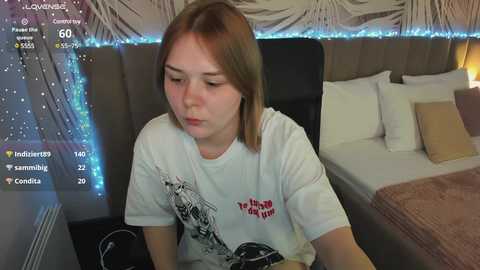 Media: Video of a young Caucasian woman with light brown hair, wearing a white t-shirt with a graphic, playing a video game in a dimly lit bedroom with a bed and string lights.