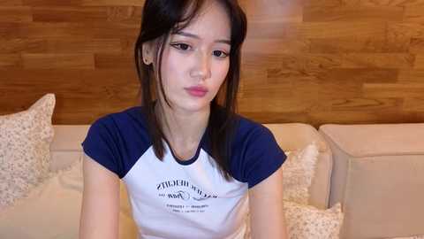 Media: Video of an Asian woman with long black hair, wearing a navy-blue and white T-shirt with a logo, sitting on a beige couch against a wooden wall backdrop.