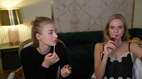 Media: Video of two women with pale skin, one with blonde hair in a bun, one with long blonde hair, both holding lollipops, in a living room with a green sofa, lamp, and abstract wall art.