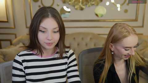 Media: Video of two young girls in a posh living room. One has straight brown hair, the other blonde. Both wear casual, striped tops. Background features ornate gold-framed mirrors, plush beige couch, and stylish decor.