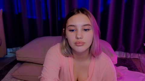 Media: Video of a young woman with light skin and shoulder-length, straight, pastel-colored hair, wearing a pink top, sitting on a bed with purple and blue lighting in the background.