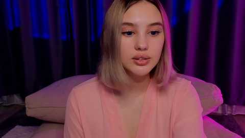 Media: Video of a young Caucasian woman with light skin, shoulder-length blonde hair, and light makeup, wearing a pink robe, sitting on a bed with purple and blue curtains in the background.