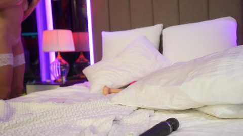 Media: Video of a messy, unmade bed with white linens, featuring a partially visible, tan-skinned person with a dildo in their hand, and a black vibrator on the bed. Purple and white neon lights illuminate the background.