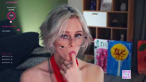 Media: A video of a blonde woman with glasses, wearing a red halter top, teasingly covering her mouth with her fingers. She is indoors, near a wooden bookshelf, with a green wall background.