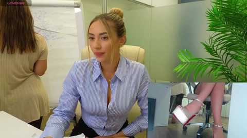 Media: Video of a blonde woman with a bun, wearing a blue and white striped blouse, seated at a desk in an office with plants and a coworker visible.