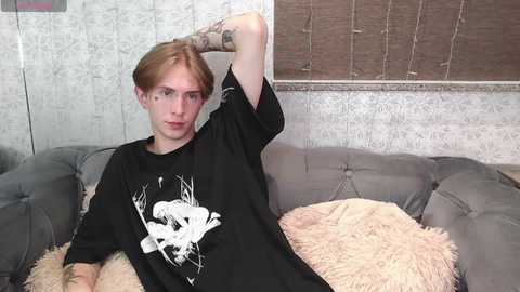 Media: Video of a young, slender, androgynous person with light skin and long, straight, reddish-brown hair, wearing a black graphic tee and lounging on a gray tufted sofa with beige throw pillows.