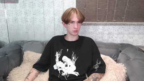 Media: Video of a young, fair-skinned, androgynous person with long, light brown hair, wearing a black T-shirt with a white graphic of a woman's face and skull, sitting on a gray, tufted sofa with beige, fluffy pillows, against a textured, light gray wall with a wooden blind.