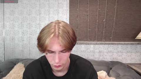 Media: Video of a young, fair-skinned man with long, light brown hair, wearing a black shirt, sitting on a gray couch with a beige blanket, against a gray, textured wall with brown blinds.