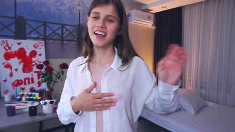 Media: Video of a young woman with long brown hair, wearing a white shirt, smiling, in a modern room with art, bed, and air conditioning.