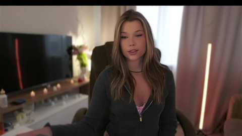 Media: A video of a young woman with long, wavy brown hair, wearing a dark jacket, sitting indoors with a flat-screen TV in the background.