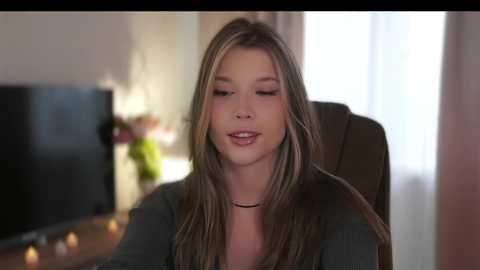 Media: A video of a young woman with long, straight blonde hair, wearing a black choker and gray top, sitting in a dimly lit room with a TV and vase of flowers in the background.