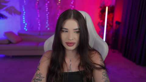 Media: Video of a tattooed woman with long dark hair, wearing a black strapless top, sitting in a white gaming chair. Background features colorful string lights and a couch, creating a vibrant, modern room ambiance.